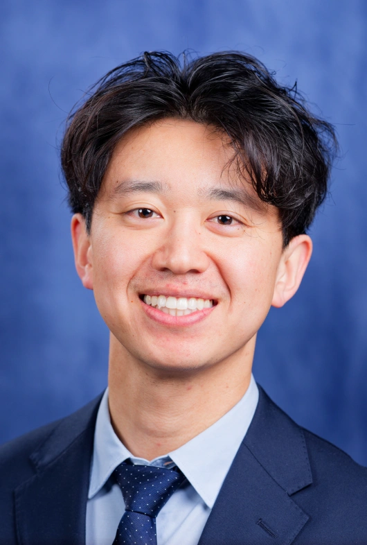 Professional headshot of smiling male dentist against blue background, wearing dark suit and tie.
