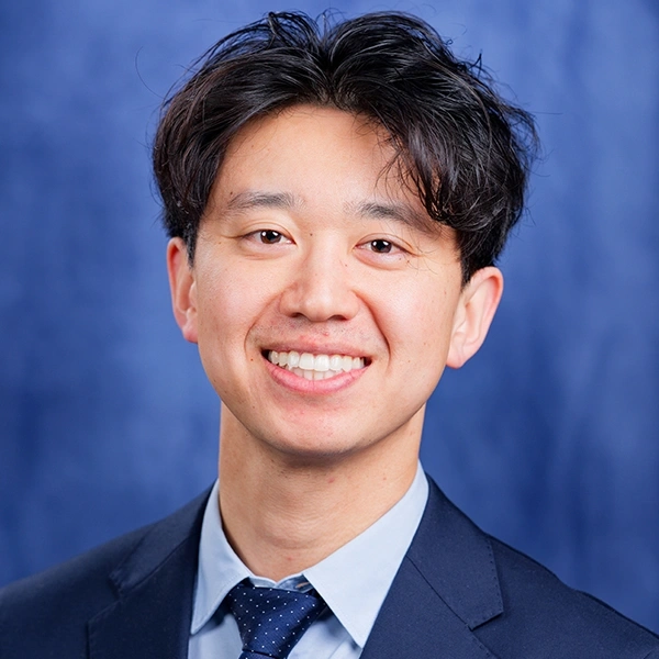 Professional headshot of smiling male dentist in dark suit and tie against blue background.