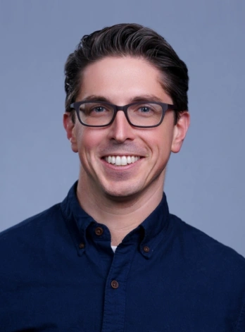 Studio headshot of a smiling man with short dark hair, wearing glasses and a dark blue button-down shirt against a solid light blue background.