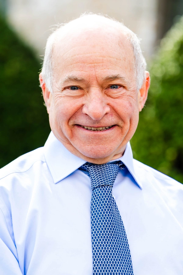 Close-up headshot of a smiling man with short white hair and blue eyes, wearing a light blue dress shirt and a blue patterned tie.