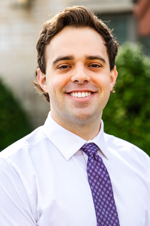 Close-up portrait of a smiling man with brown wavy hair, wearing a white dress shirt and a patterned purple tie.
