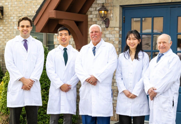 Group portrait of five dental professionals wearing white lab coats, standing together and smiling in front of a brick building with a dark blue door.