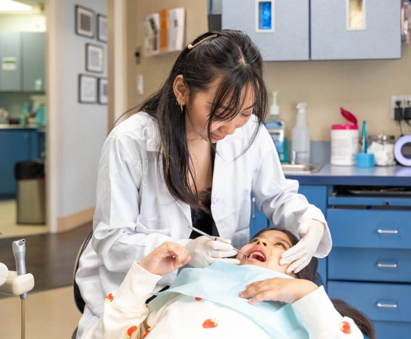 A female dentist wearing a white lab coat and dental gloves performing a checkup on a young child lying back in a dental chair.