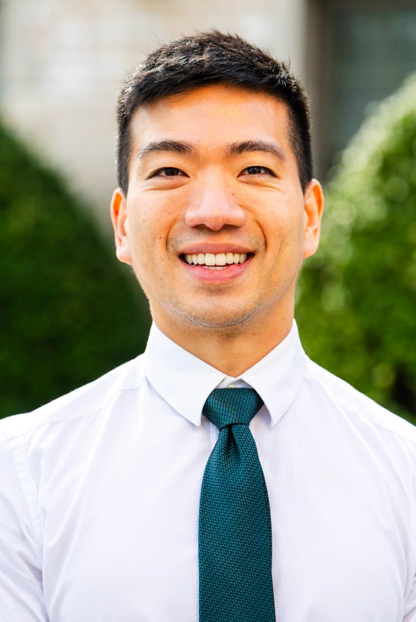 Close-up outdoor headshot of a smiling man with short dark hair, wearing a white button-down shirt and a dark green tie.