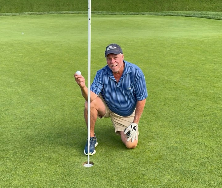 A man in a blue striped polo, shorts, and a UCSB baseball cap kneeling on a golf green holding up a golf ball next to the flagstick hole.