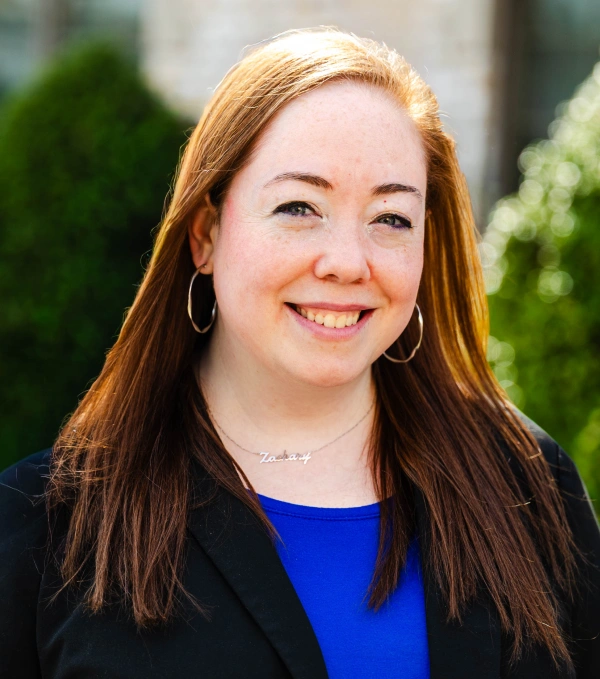 Outdoor headshot of a smiling woman with long straight auburn hair, hoop earrings, and a name necklace, wearing a dark blazer over a blue top.