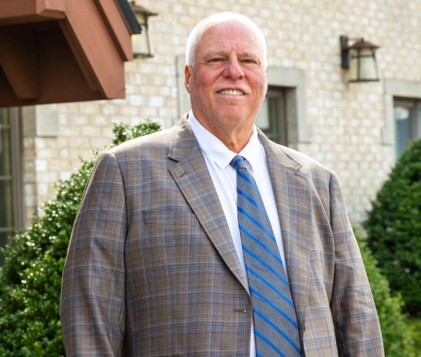 Medium shot of a smiling man with short white hair, wearing a plaid suit jacket, white dress shirt, and a blue diagonally striped tie, standing outdoors in front of a brick building.