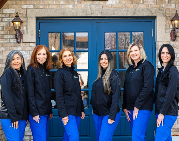 Six women wearing black zip-up jackets over royal blue scrub pants standing outside in a row against a brick building and blue double doors.