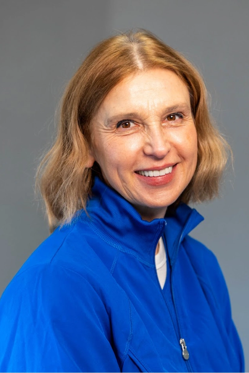 Studio headshot of a smiling woman with short, wavy blonde-and-brown hair, wearing a bright blue zip-up jacket against a plain gray background.