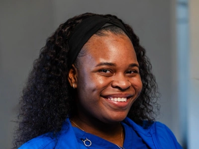 Close-up studio headshot of a woman with long, curly dark hair wearing a black thick headband and a bright blue top, smiling widely toward the camera.