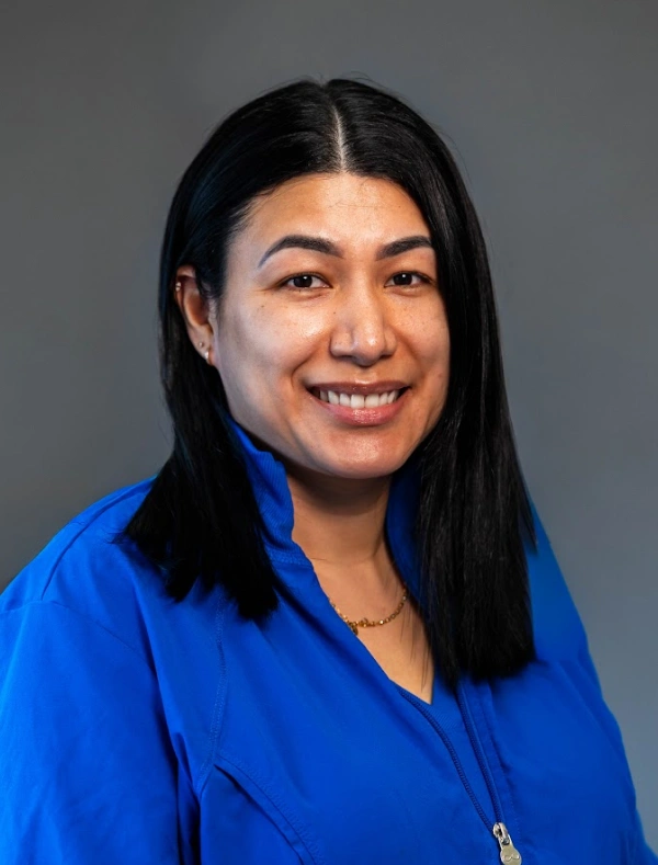 Portrait of a woman with straight shoulder-length black hair wearing a bright blue jacket and a gold necklace against a dark gray backdrop.