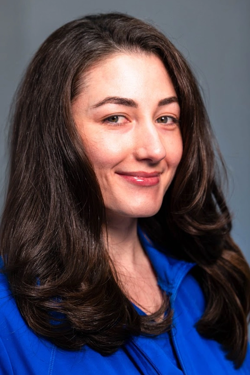 Headshot of a woman with long, dark brown hair and green eyes wearing a bright blue jacket against a solid gray backdrop, smiling gently at the camera.