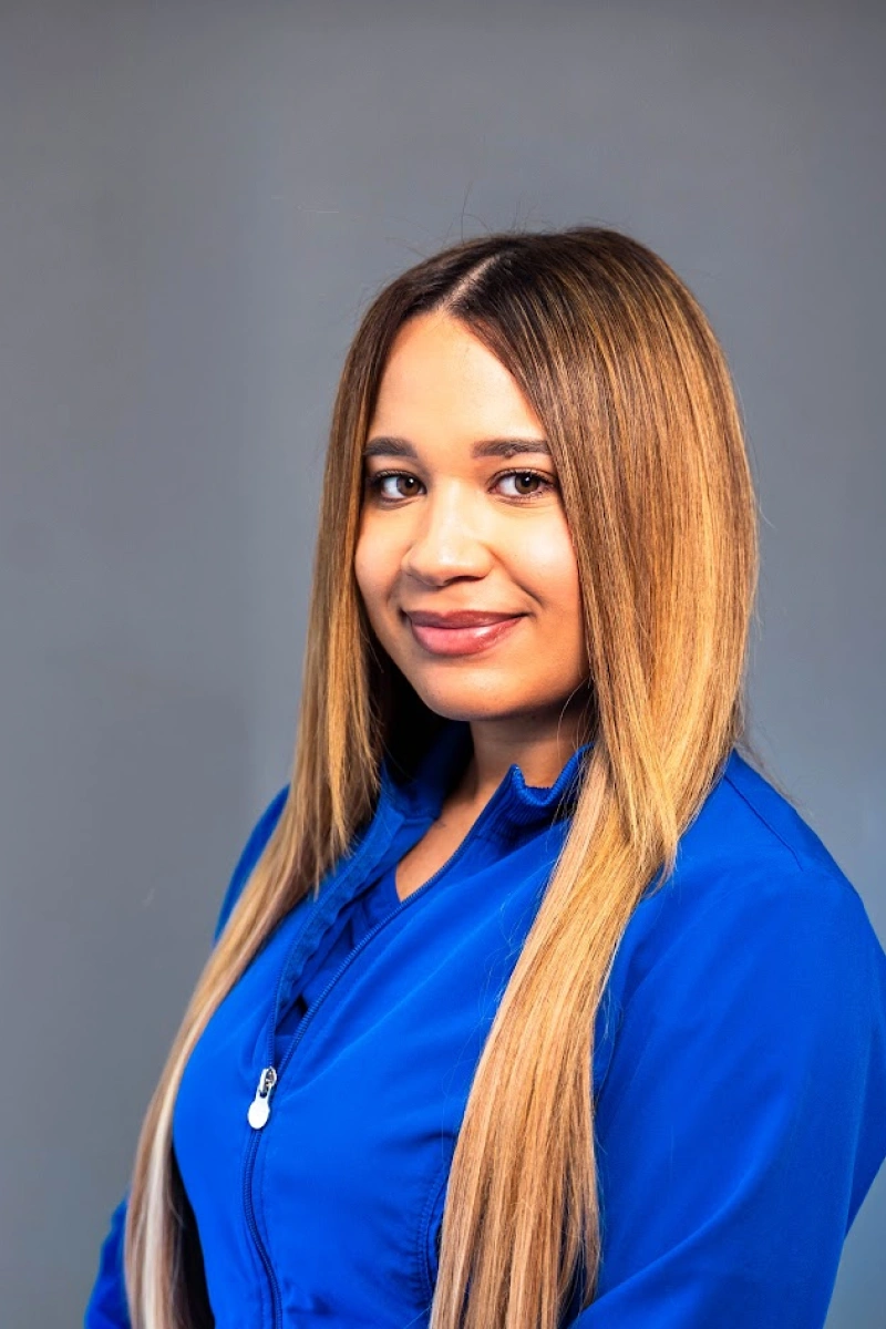 Studio portrait of a woman with straight, long blonde hair with darker roots, wearing a bright blue zip-up jacket against a neutral gray background.