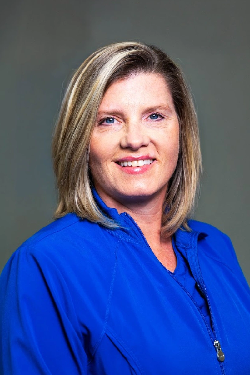 Studio headshot of a smiling woman with short blonde hair with dark lowlights, wearing a bright blue jacket against a gray backdrop.