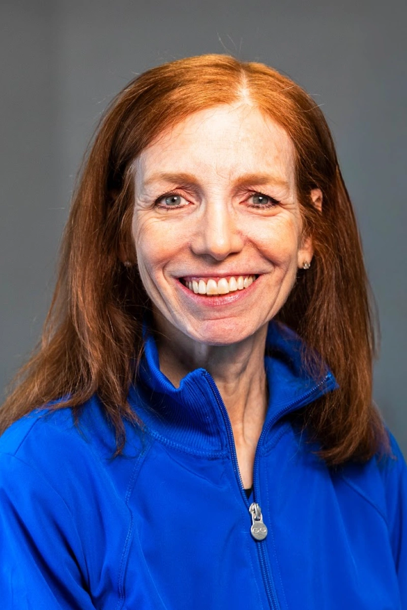 Close-up studio headshot of a smiling woman with long reddish-brown hair, wearing a bright blue zip-up jacket against a smooth gray backdrop.