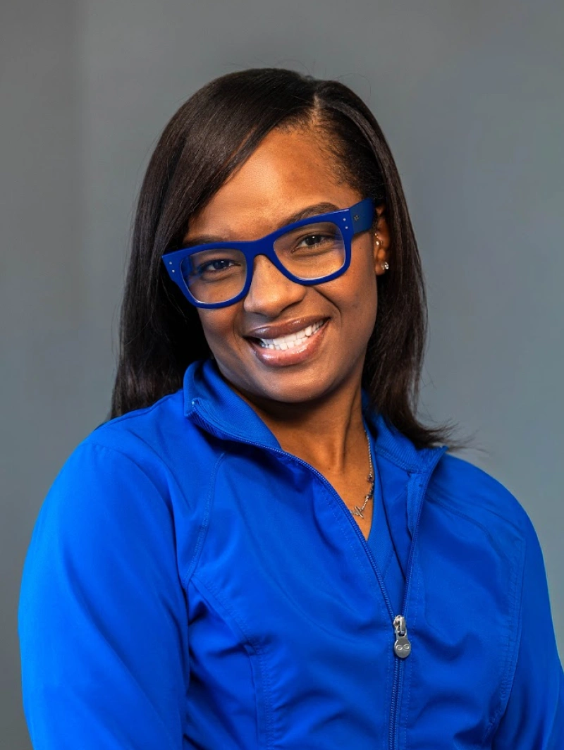 Headshot of a smiling woman with dark straight hair, wearing bright blue glasses frames and a matching bright blue jacket.