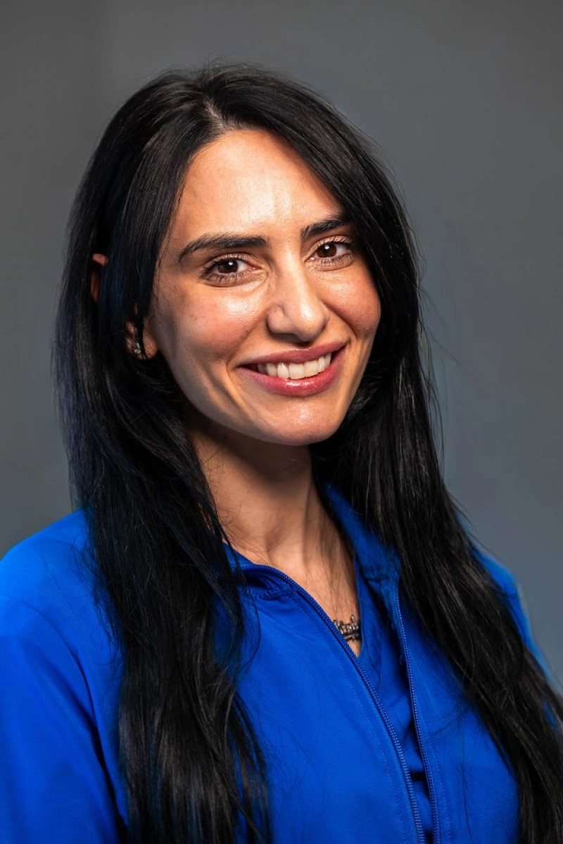 Studio portrait of a smiling woman with long, straight black hair wearing a bright blue jacket against a solid dark gray backdrop.