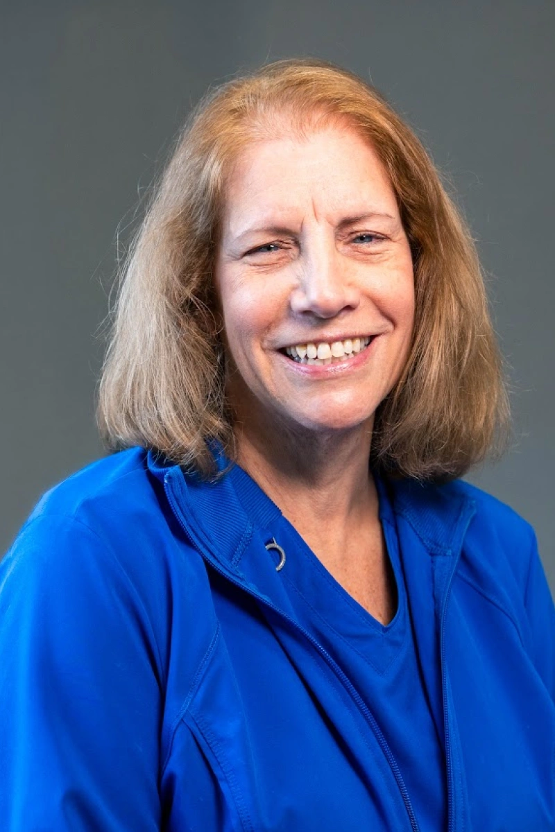 Studio headshot of a woman with shoulder-length dirty blonde hair and blue eyes, wearing a bright blue scrub top and smiling directly at the camera.