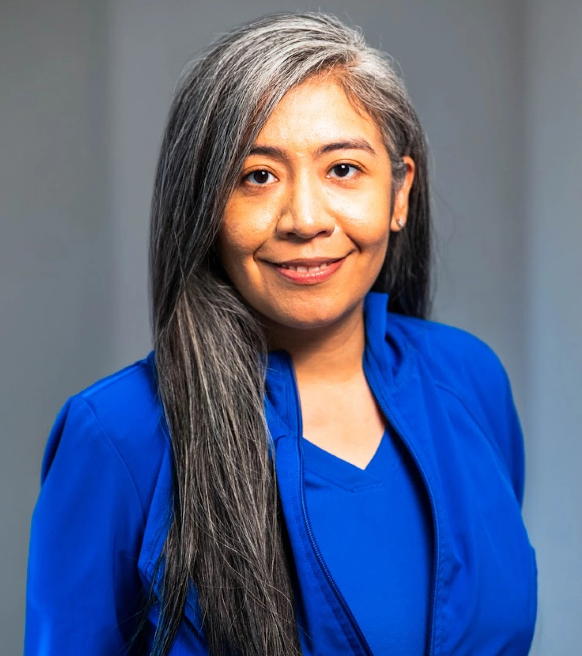 Headshot of a woman with long, side-swept dark hair mixed with gray strands, wearing a bright blue zip-up jacket and smiling softly against a gray background.