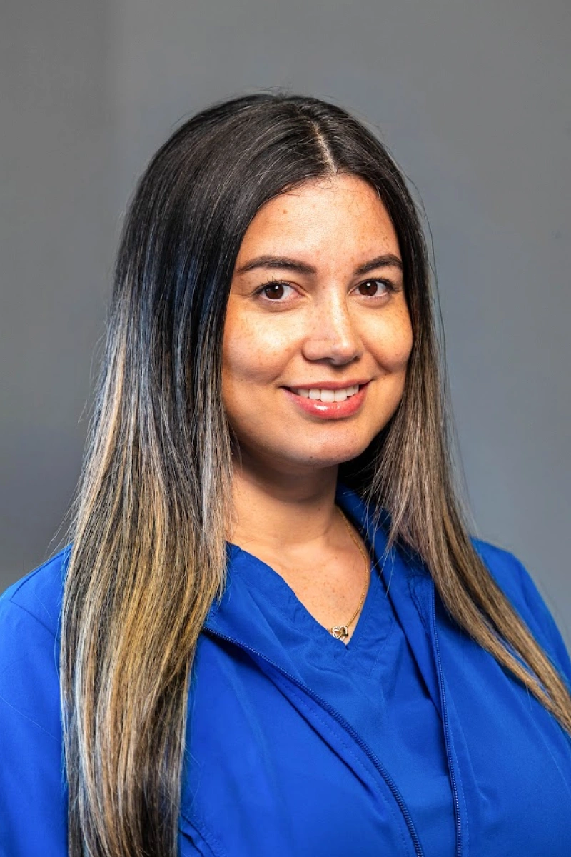 Studio portrait of a smiling woman with long, straight dark hair featuring light blonde highlights, wearing a bright blue jacket against a neutral gray background.