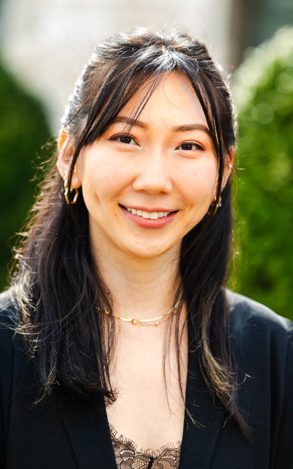 Close-up outdoor headshot of a smiling woman with long dark hair, wearing a black blazer and gold jewelry against a green backdrop.