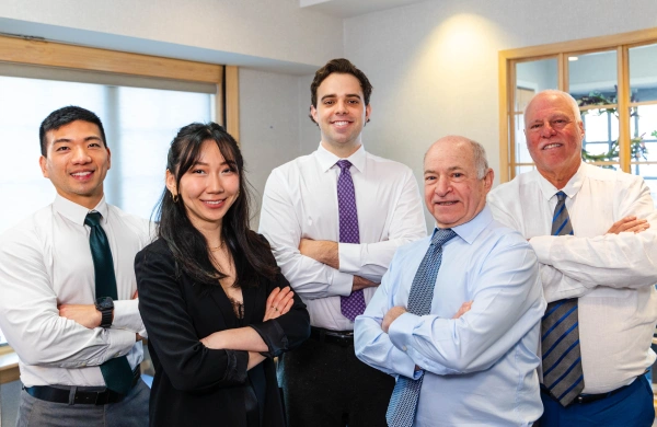 Five business professionals standing indoors with their arms crossed, smiling at the camera in professional attire.