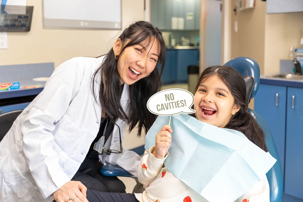 A smiling female dentist in a white coat leans over a young girl sitting in a blue dental chair. The girl is holding a speech bubble prop that reads "NO CAVITIES!" and smiling happily.
