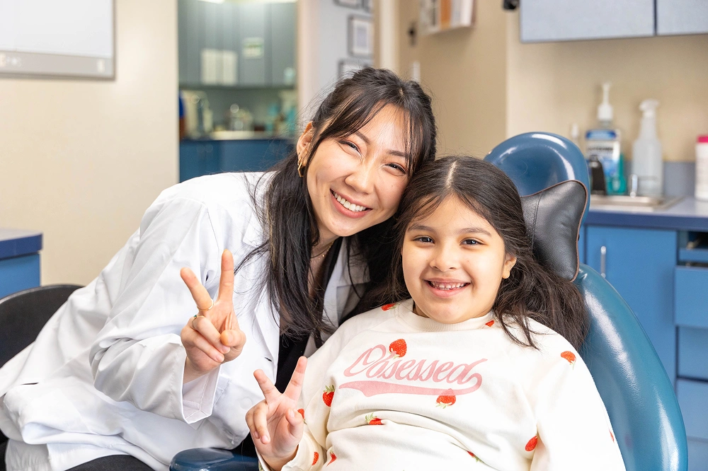 The female dentist and the young girl pose closely together in the dental office, both smiling and making peace signs with their hands while the girl sits in the blue dental chair.